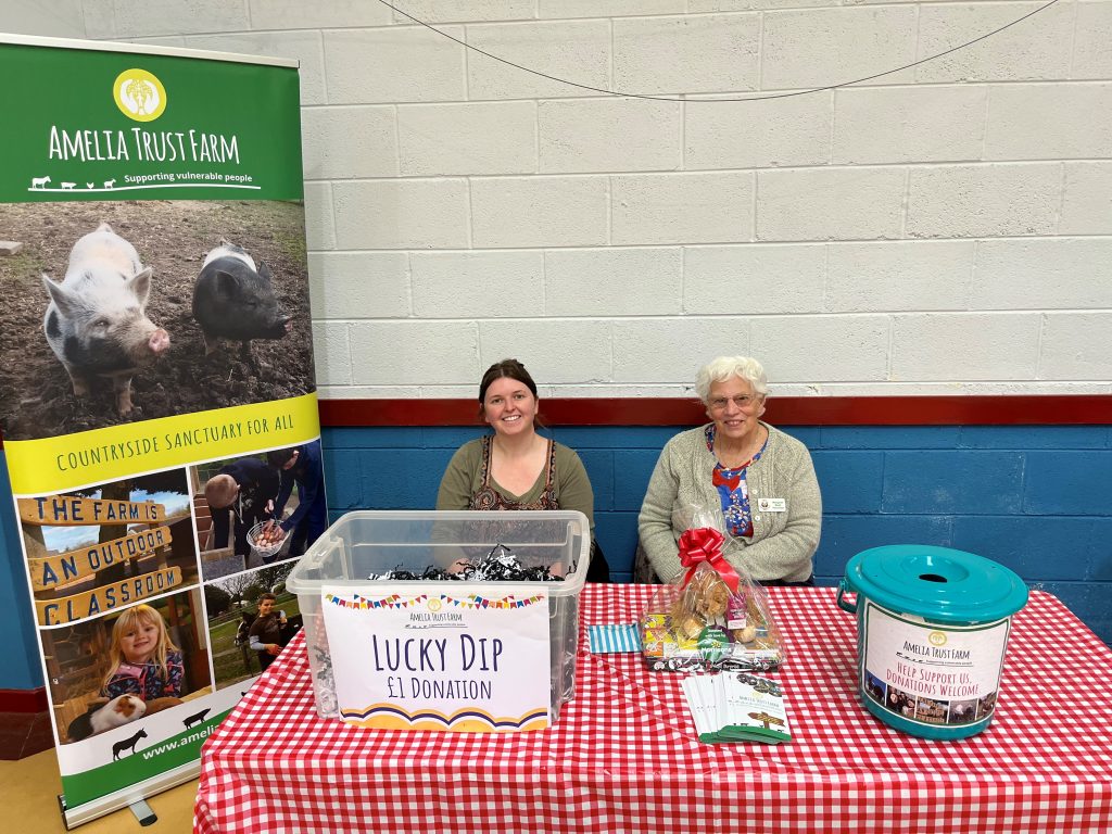 Two volunteers sitting behind a red check tablecloth with a lucky dip and donation bucket.